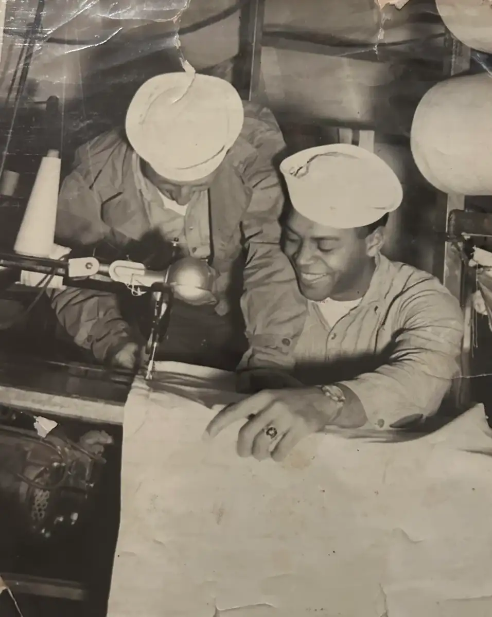 Two sailors in uniform work together at a sewing machine, one guiding fabric while smiling and the other focused on the task. The scene appears in black and white, suggesting it is from a past era.