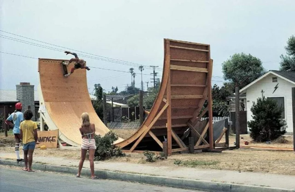 A skateboarder rides on a large wooden half-pipe in a suburban yard, watched by three people standing nearby. There are houses, trees, and power lines in the background.