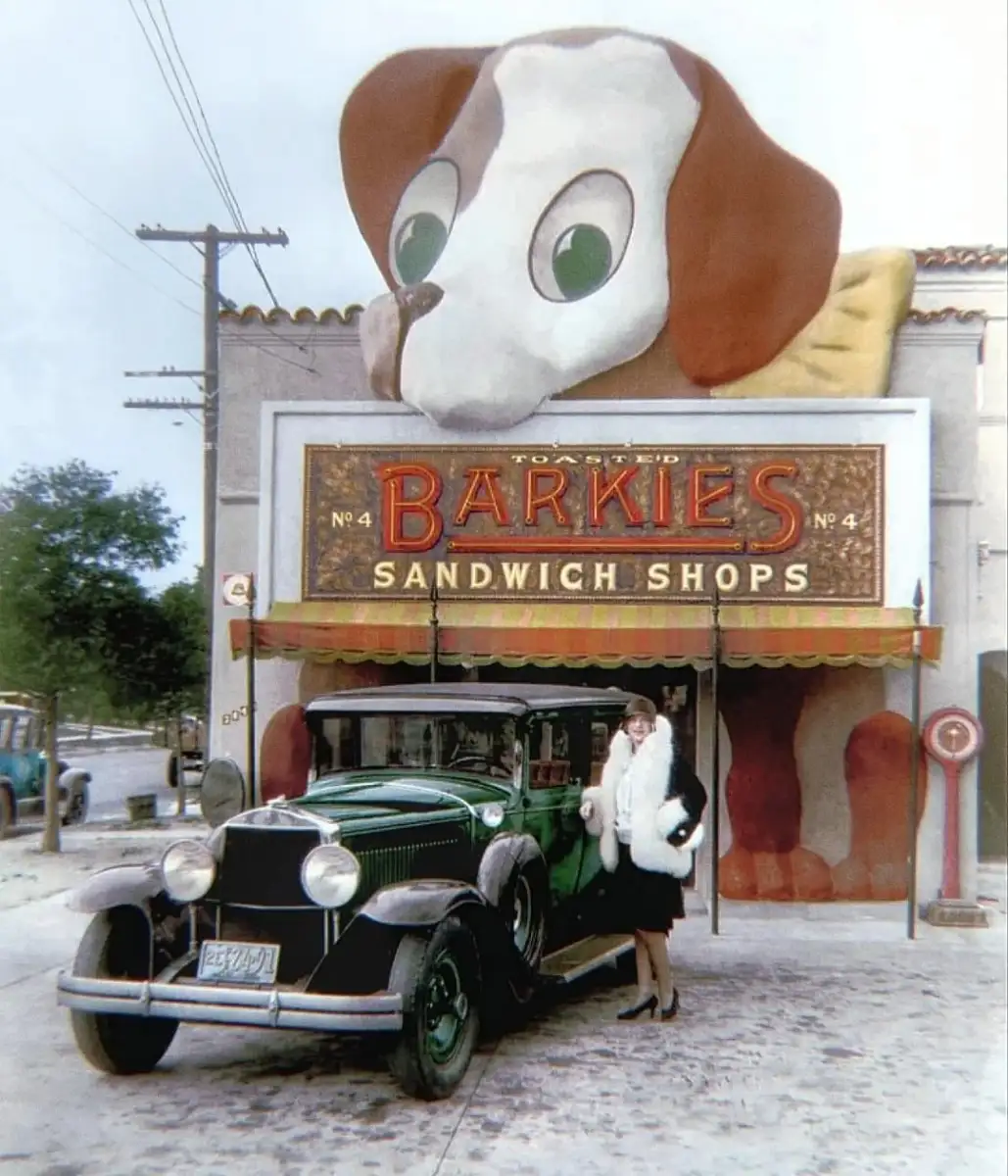 A woman in a fur-trimmed coat stands next to a vintage car in front of Barkies Sandwich Shop, whose building features a giant cartoon dog head and paws above the entrance.