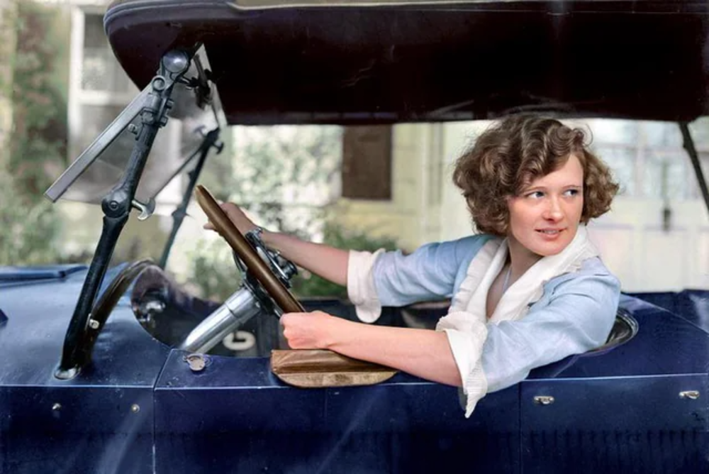 A young woman with curly hair sits in a vintage blue convertible car, looking back over her shoulder and smiling while holding the steering wheel, dressed in a light blouse with rolled sleeves.
