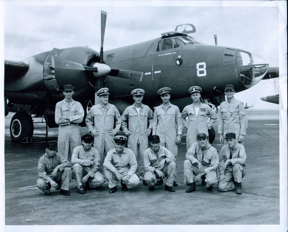 Black-and-white photo of thirteen uniformed men posing in front of a large military airplane marked with the number 8, on an airfield. Some are standing, others are kneeling, all looking at the camera.