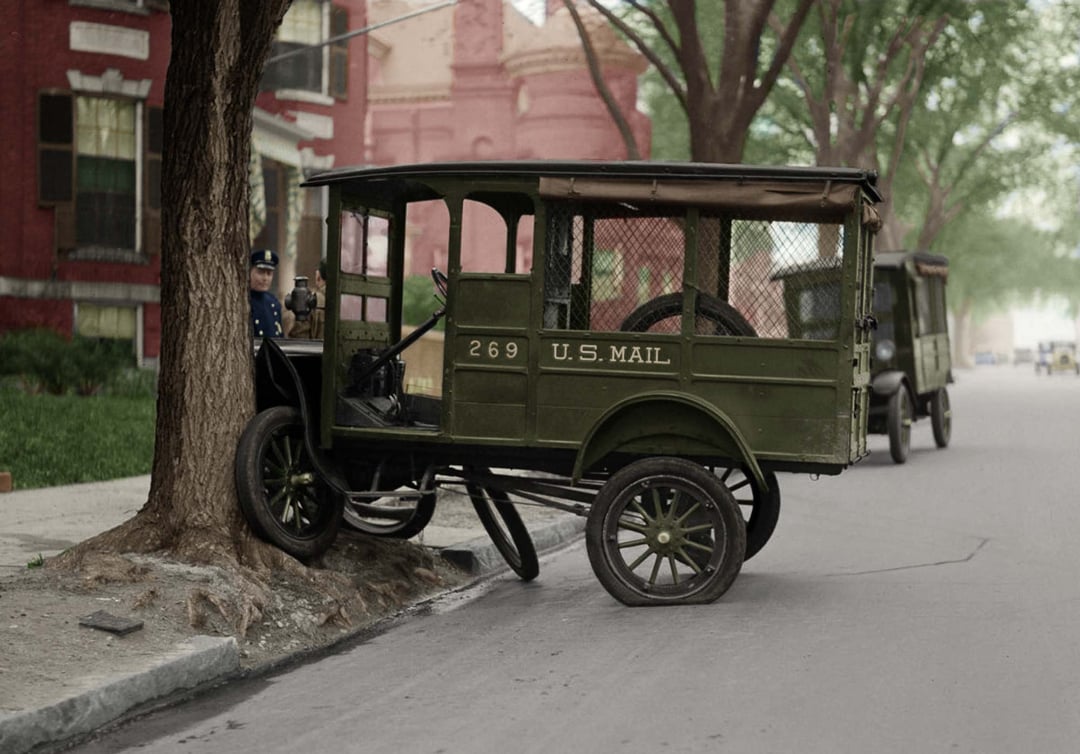 A vintage green U.S. Mail truck, number 269, has crashed into a tree on a city street, with the front wheels lifted off the ground. A uniformed person stands nearby. Another mail truck is parked further down the road.