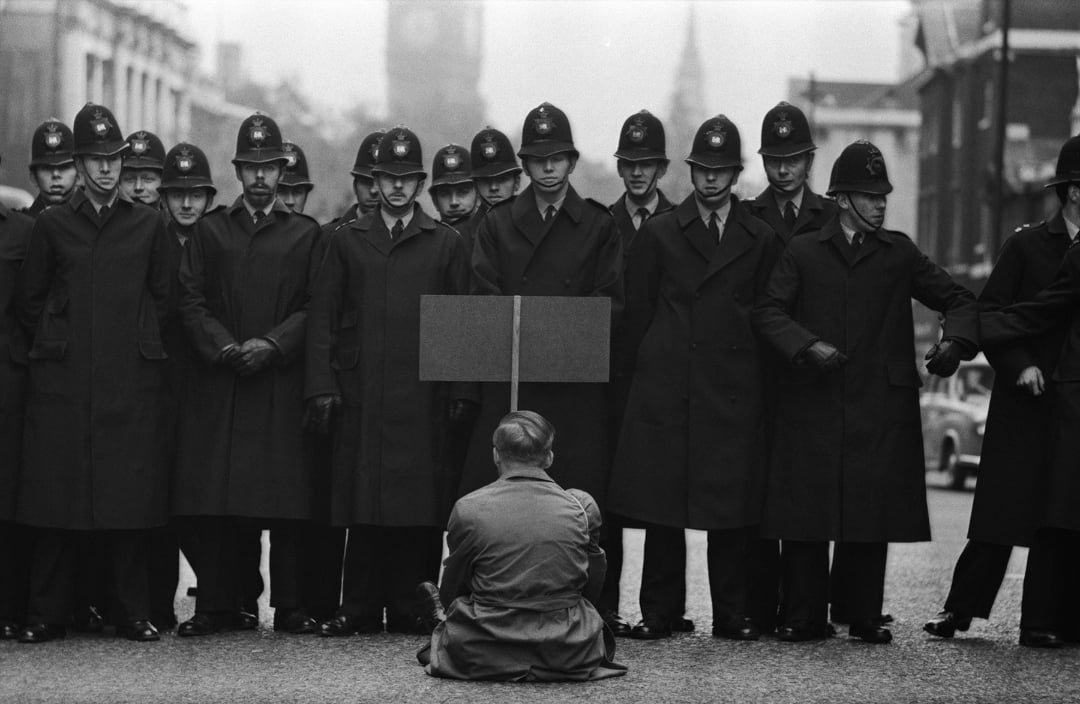 A person sits on the ground facing a line of police officers in uniform and helmets, holding a blank sign in protest on a city street. Buildings and a clock tower are visible in the background.