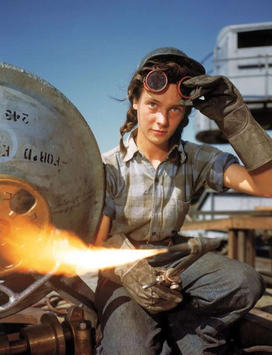 A young woman wearing gloves, goggles, and work clothes uses a blowtorch, with a bright flame visible. She sits outdoors, possibly at an industrial or construction site, looking directly at the camera.