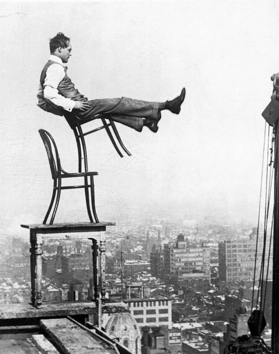 A man in a suit balances precariously on a chair that is tilted on the back legs, which is stacked on another chair and table, high above a city skyline with buildings in the background.
