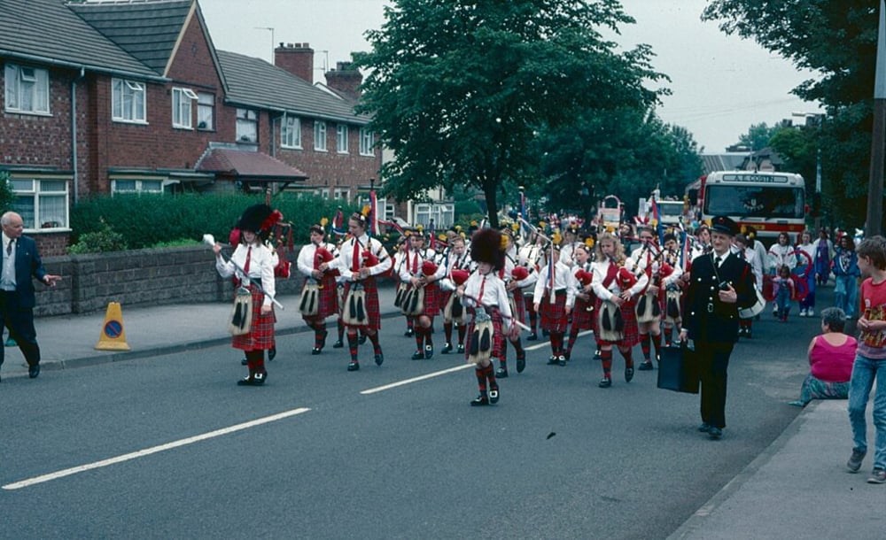 A marching band in traditional Scottish attire, including kilts and bagpipes, parades down a residential street lined with houses and onlookers; a bus and more spectators are visible in the background.