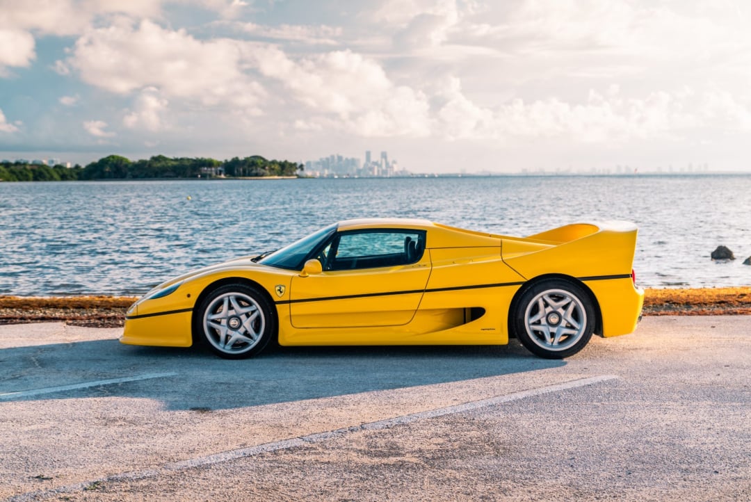 A bright yellow Ferrari sports car is parked beside a waterfront, with a city skyline visible in the distance under a partly cloudy sky.