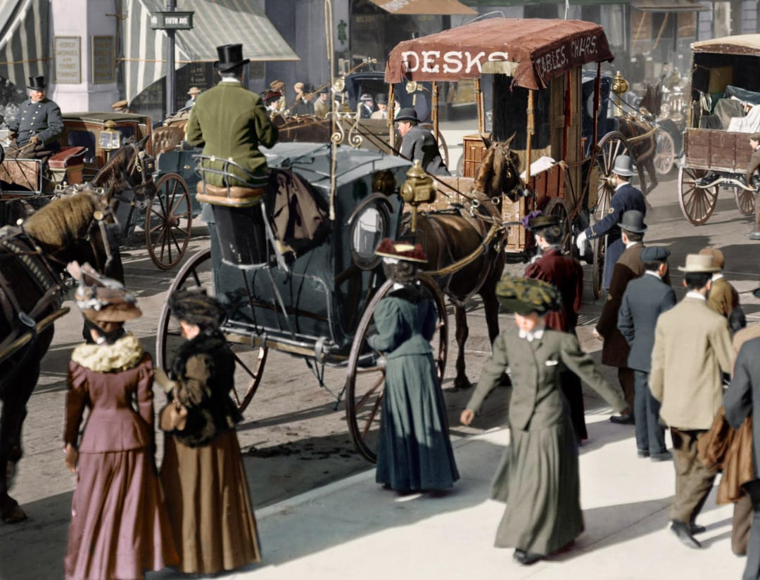 A busy city street scene from the early 1900s, with people in period clothing walking and horse-drawn carriages on the road. Signs and storefronts are visible in the background.