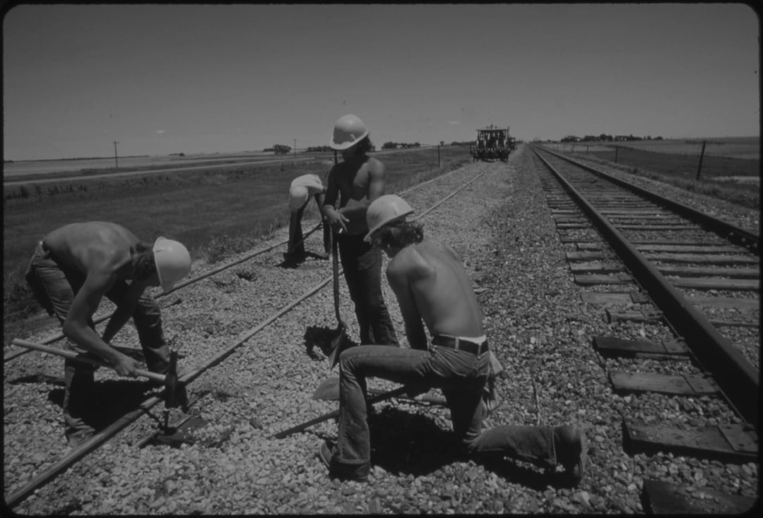 Four shirtless workers wearing hard hats repair a railroad track in a rural area under a clear sky. Two kneel using tools on the rail while others stand nearby. Train tracks and open fields stretch into the distance.
