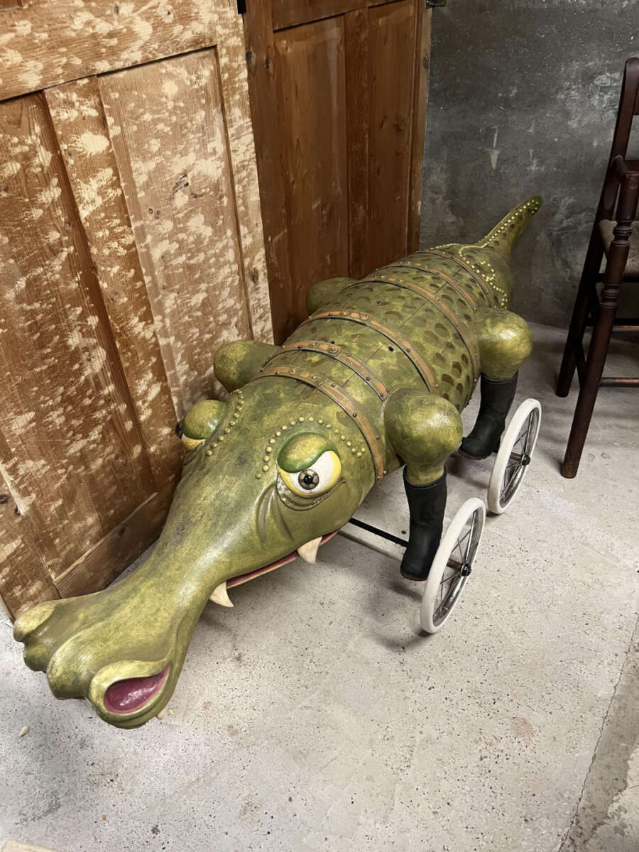 A whimsical, green, crocodile-shaped ride-on toy with wheels stands on a concrete floor beside a wooden cabinet. The crocodile has large eyes, white fangs, and a playful expression.