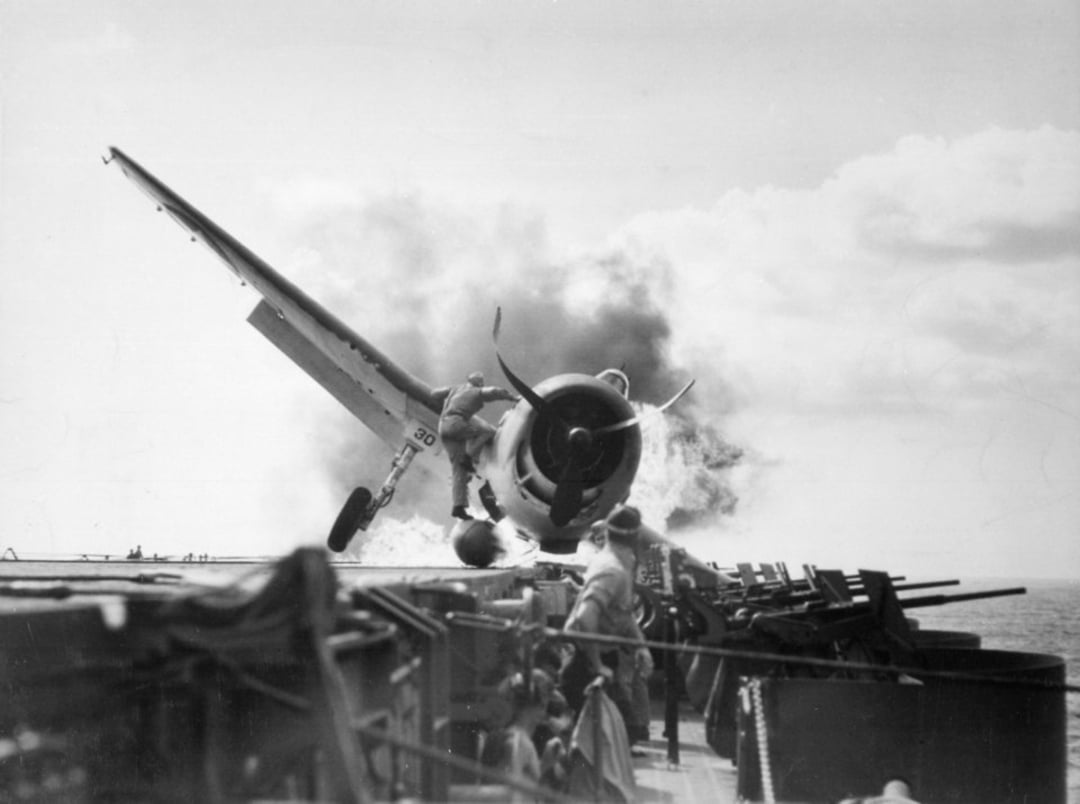 A fighter plane crashes onto an aircraft carrier deck, smoke rising from its engine as crew members scramble nearby, some watching and others ducking for cover under a sunny, partly cloudy sky.