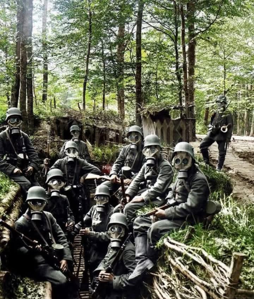 A group of soldiers in World War I uniforms and gas masks sit and stand in a trench surrounded by trees and foliage in a forested area. One soldier stands guard on the path above.