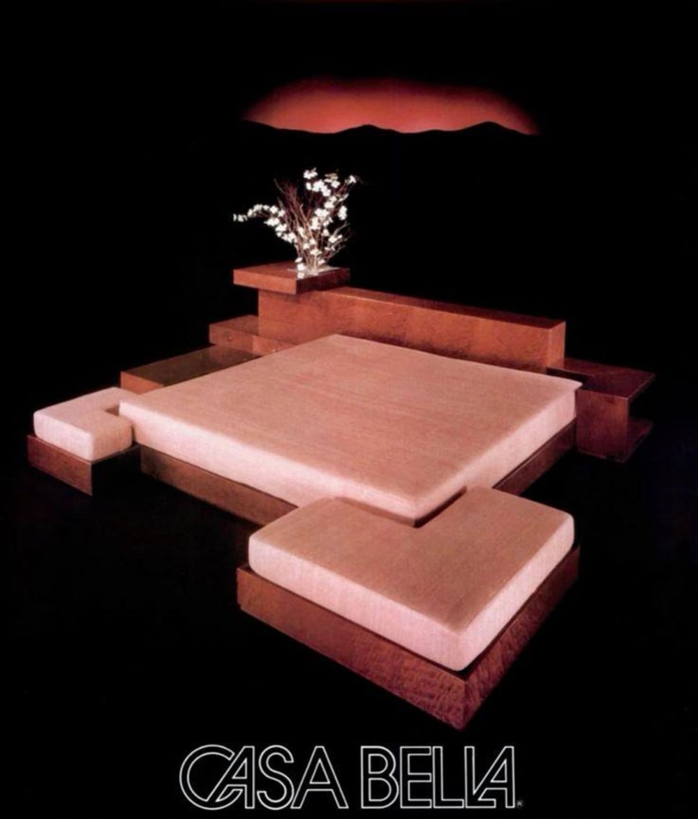 A modern, minimalist bed with beige cushions and wood platform features, accented by a low shelf with a vase of white flowers. The background is black with a subtle mountain silhouette. "CASA BELLA" is written at the bottom.