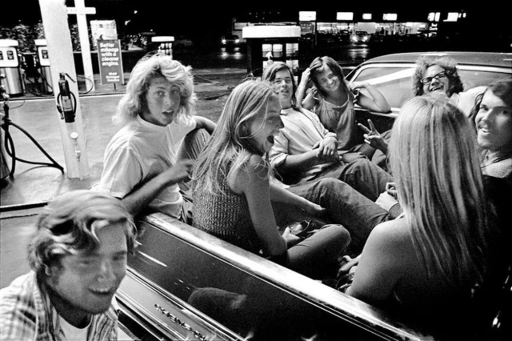 A group of young people sit and laugh together in the back of a convertible car at a gas station at night, with bright lights and gas pumps visible in the background.