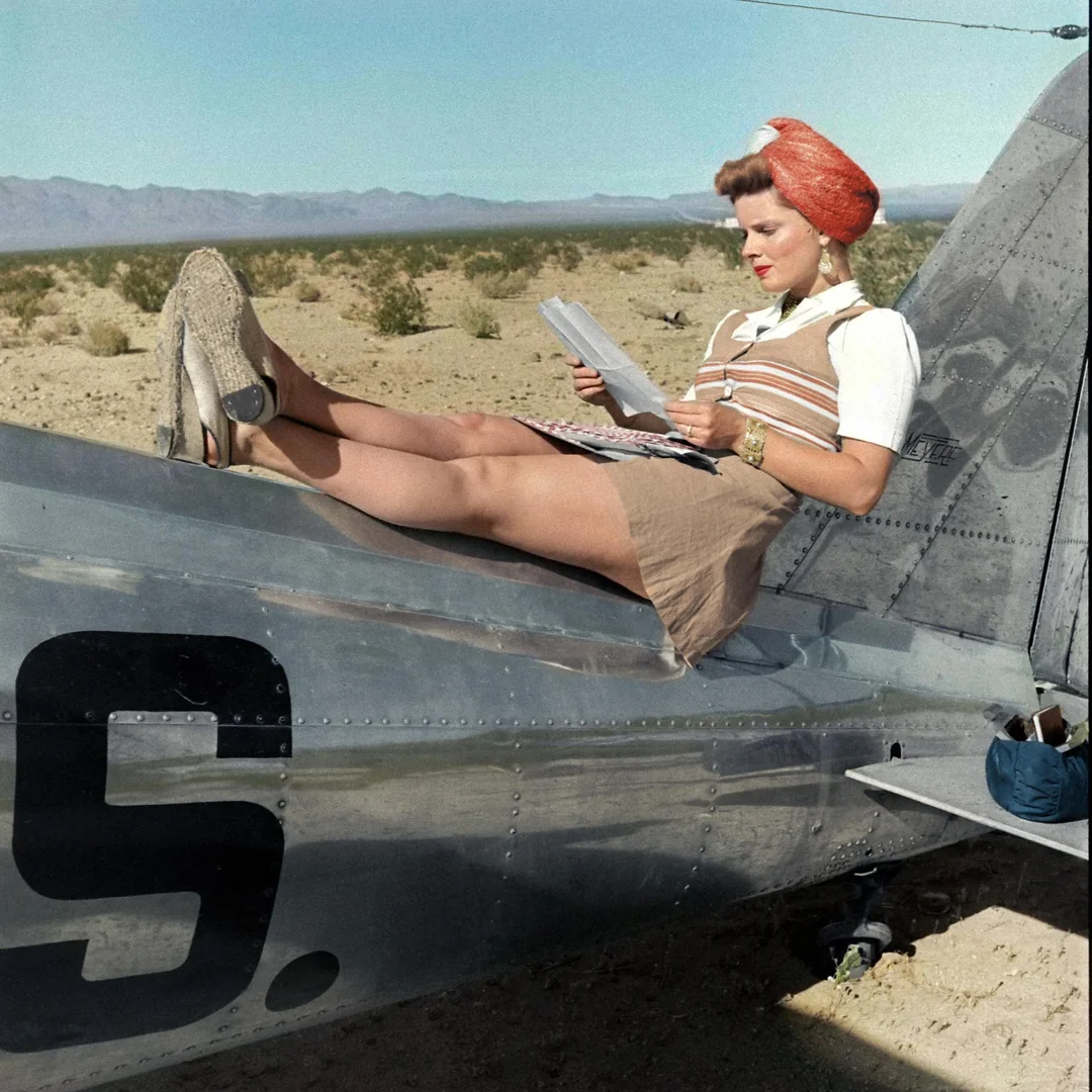 A woman in 1940s attire sits on the tail of a shiny metal airplane, reading papers in a desert landscape. She wears wedge shoes, a skirt, a blouse, and a red hat, with mountains visible in the background.