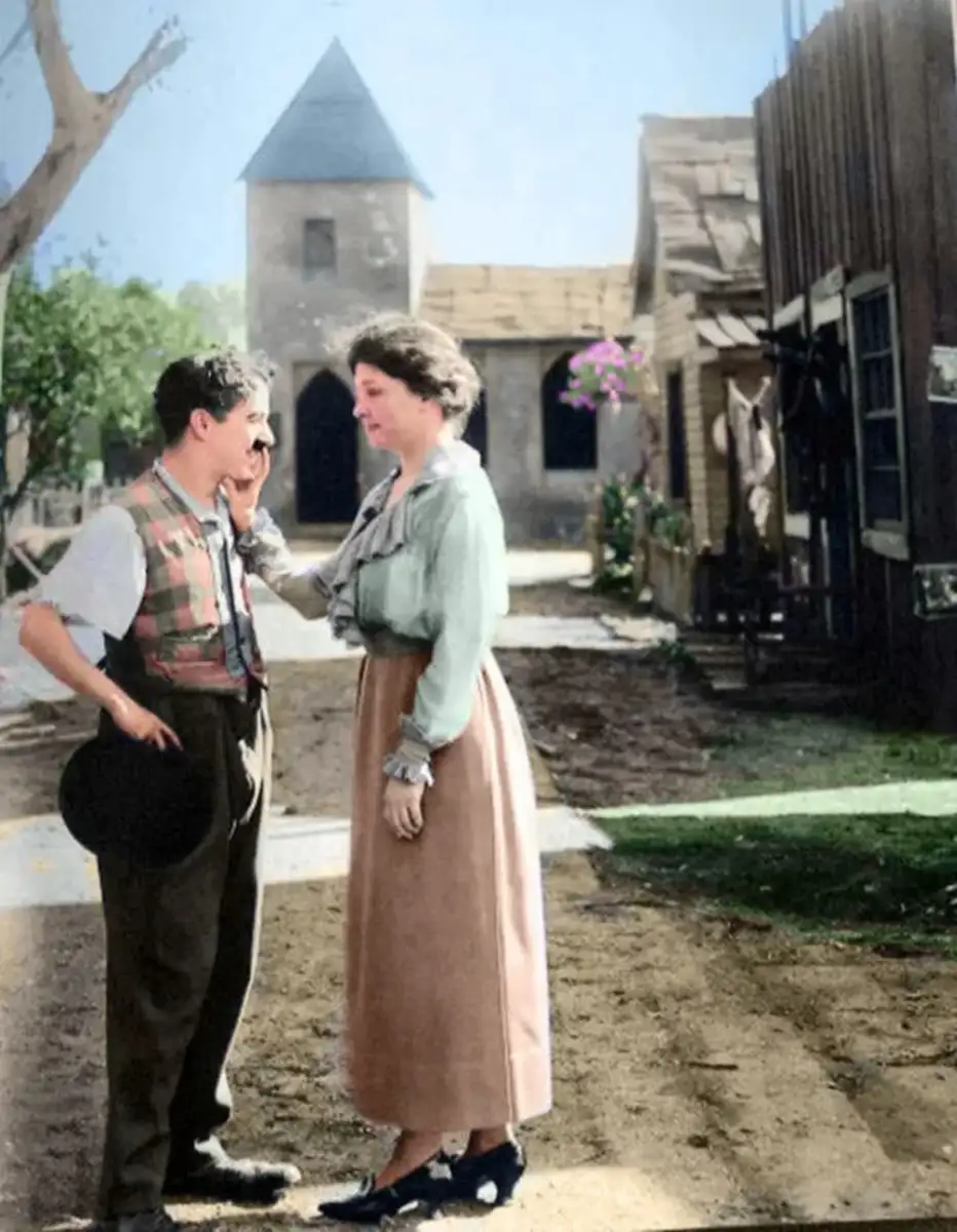 A man in vintage clothing stands holding a hat, while a woman gently touches his face. They are outside on a dirt road lined with old wooden buildings and a tower in the background, evoking an early 20th-century setting.