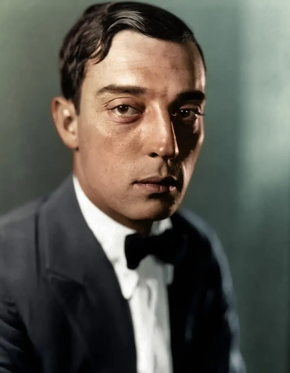 A man with short dark hair, wearing a black suit, white shirt, and black bow tie, looks directly at the camera with a serious expression against a blurred green background.