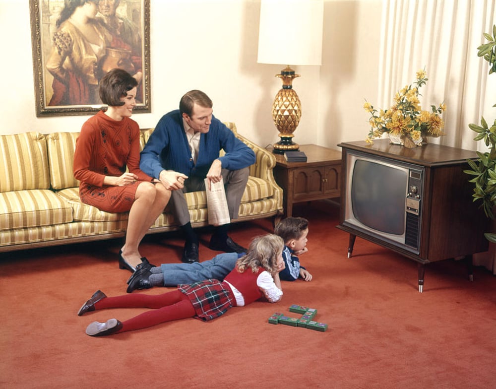 A family of four in vintage clothing watches TV in a living room. Two adults sit on a striped couch while two children lie on a red carpet facing an old-fashioned television set. A painting, lamp, and flowers decorate the room.