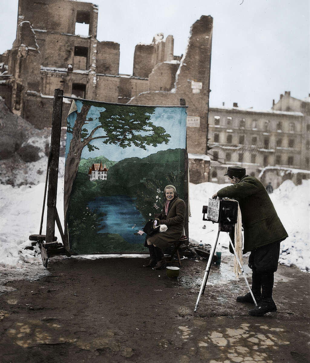A woman poses for a photograph in front of a painted backdrop of a lake and house, while a photographer takes her picture, with snowy ruins and damaged buildings in the background.