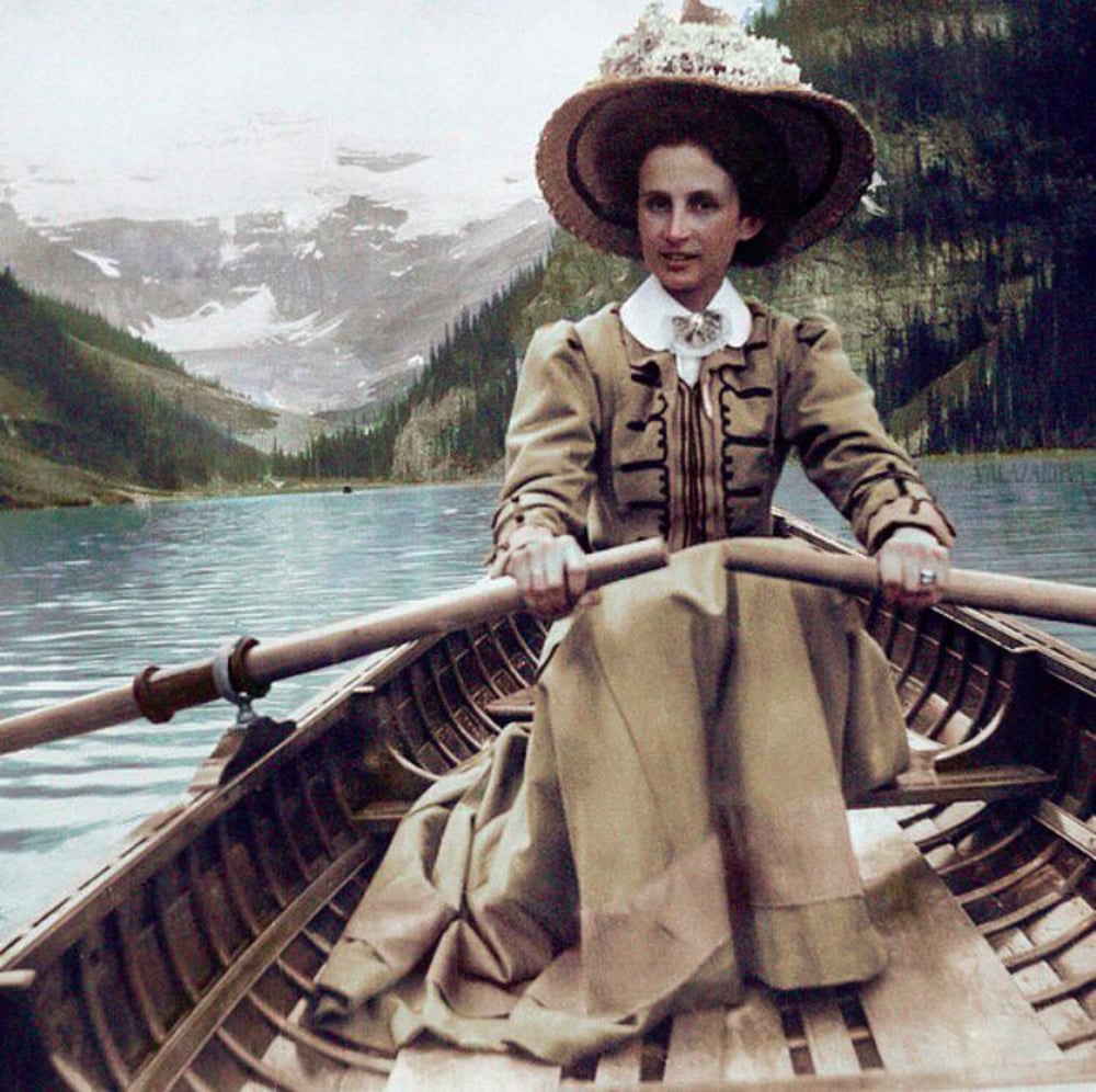 A woman in a long, vintage dress and wide-brimmed hat is rowing a wooden boat on a lake, surrounded by pine trees and snow-capped mountains in the background.