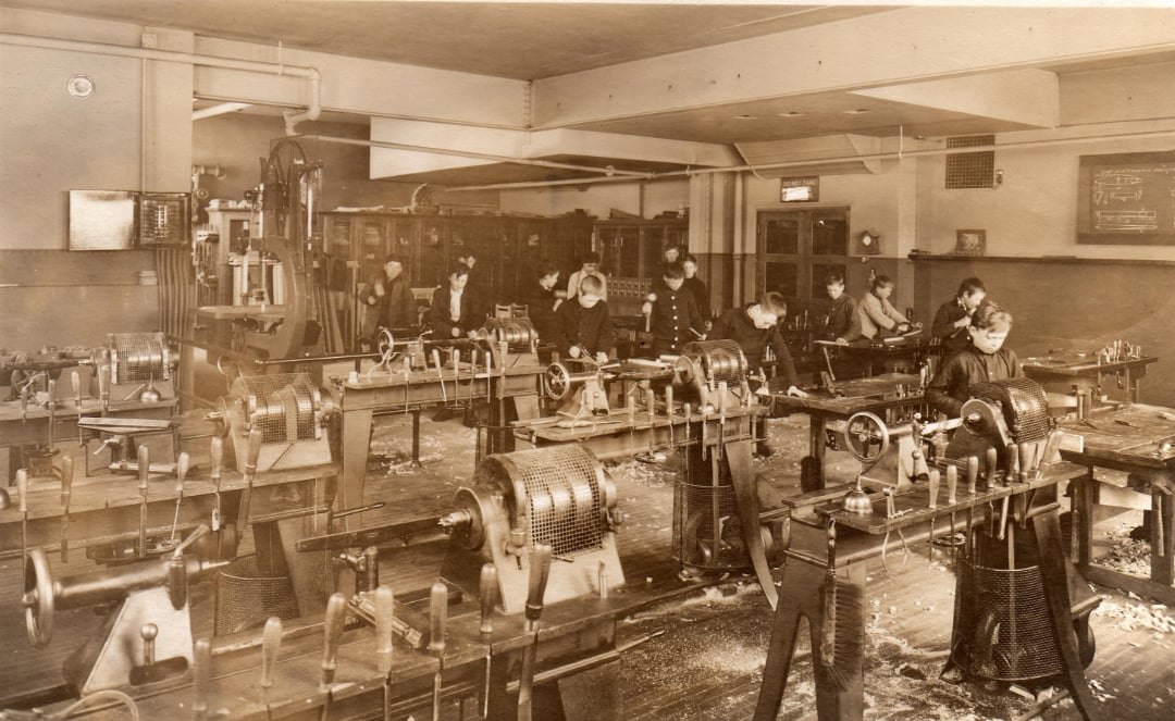 A vintage photo of people working at various machines in an early 20th-century industrial workshop or factory, with wooden floors, large equipment, and workers focused on their tasks.