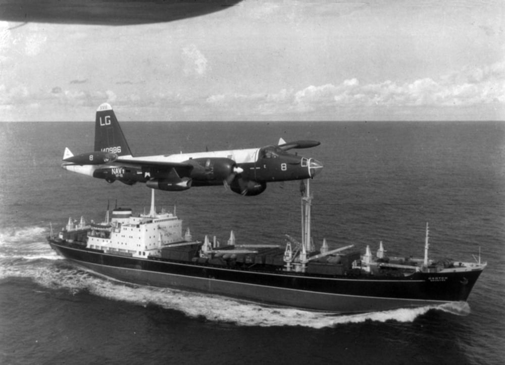 A military aircraft flies low over a cargo ship traveling at sea, with the ship creating a wake in the water. The sky above has scattered clouds.
