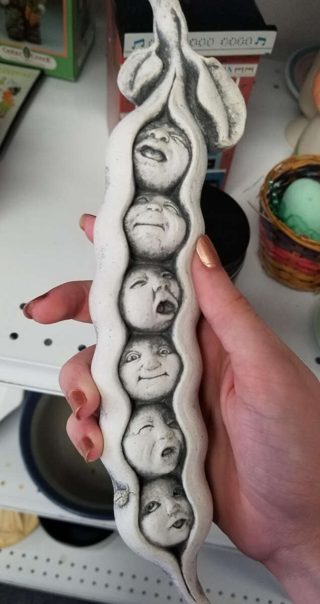 A hand holds a white ceramic sculpture shaped like a pea pod, with five peas displaying detailed, expressive human faces, each showing different emotions. The background shows shelves with various items.