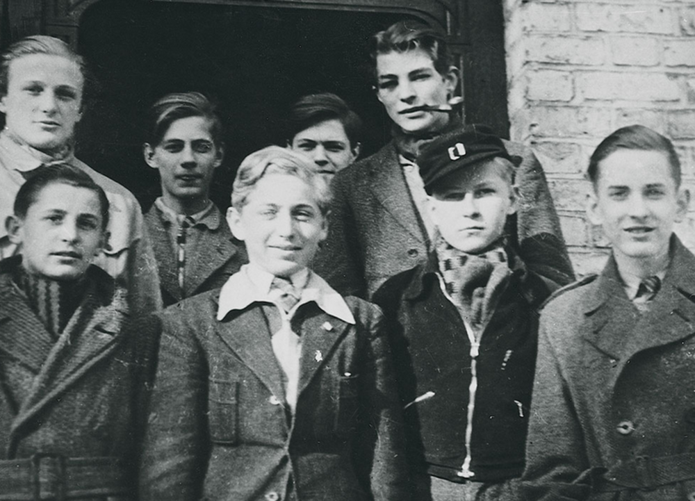 A black-and-white photo of eight teenagers standing close together outside a brick building, wearing jackets and scarves, some smiling and others with neutral expressions.
