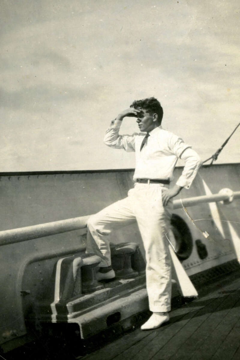 A man in light-colored clothing stands on the deck of a ship, one foot propped up, shading his eyes with his hand as he looks into the distance under a partly cloudy sky.
