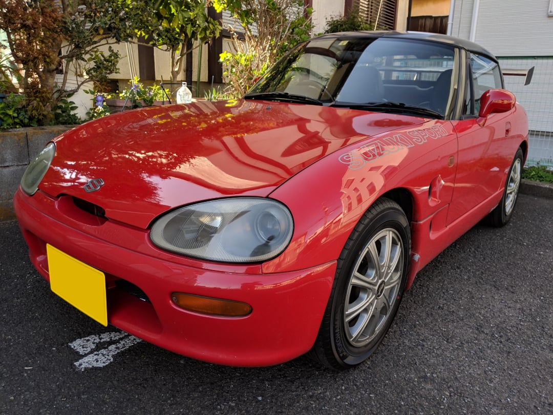 A red Suzuki Cappuccino sports car with a yellow license plate is parked on a street near greenery and buildings, reflecting sunlight on its glossy hood.
