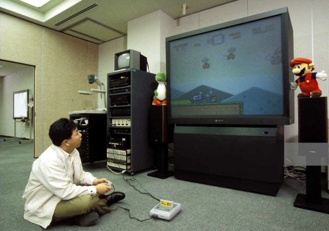A person sits cross-legged on the floor playing a Super Nintendo game on a large TV in an office, with Yoshi and Mario plush toys placed on top of the TV.