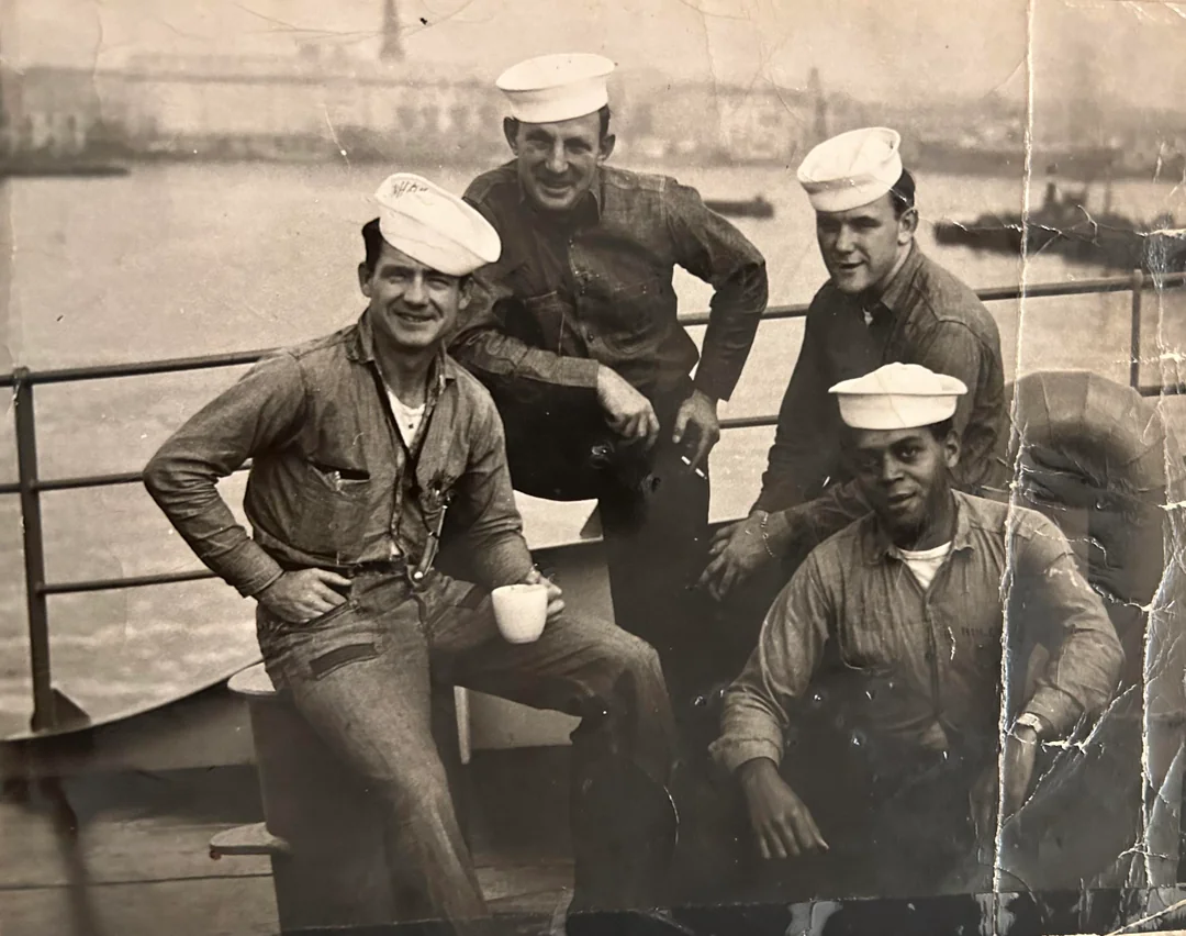 Four sailors in uniform, wearing white caps and buttoned shirts, sit and pose together on a ship’s deck. One holds a mug. The background shows water and an industrial shoreline. The photo is black and white and appears vintage.