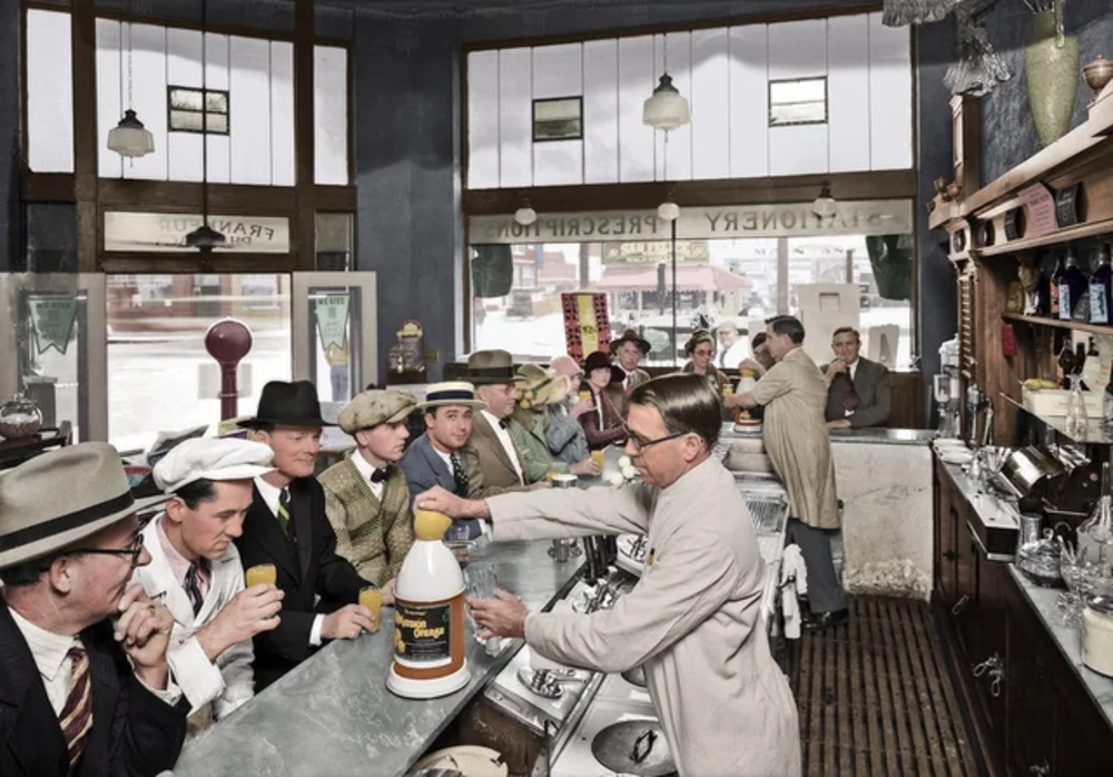 A vintage soda fountain scene shows men in suits and hats sitting at a counter, drinking and talking, while a server in a white coat prepares drinks behind the counter. The setting appears to be from the early 20th century.