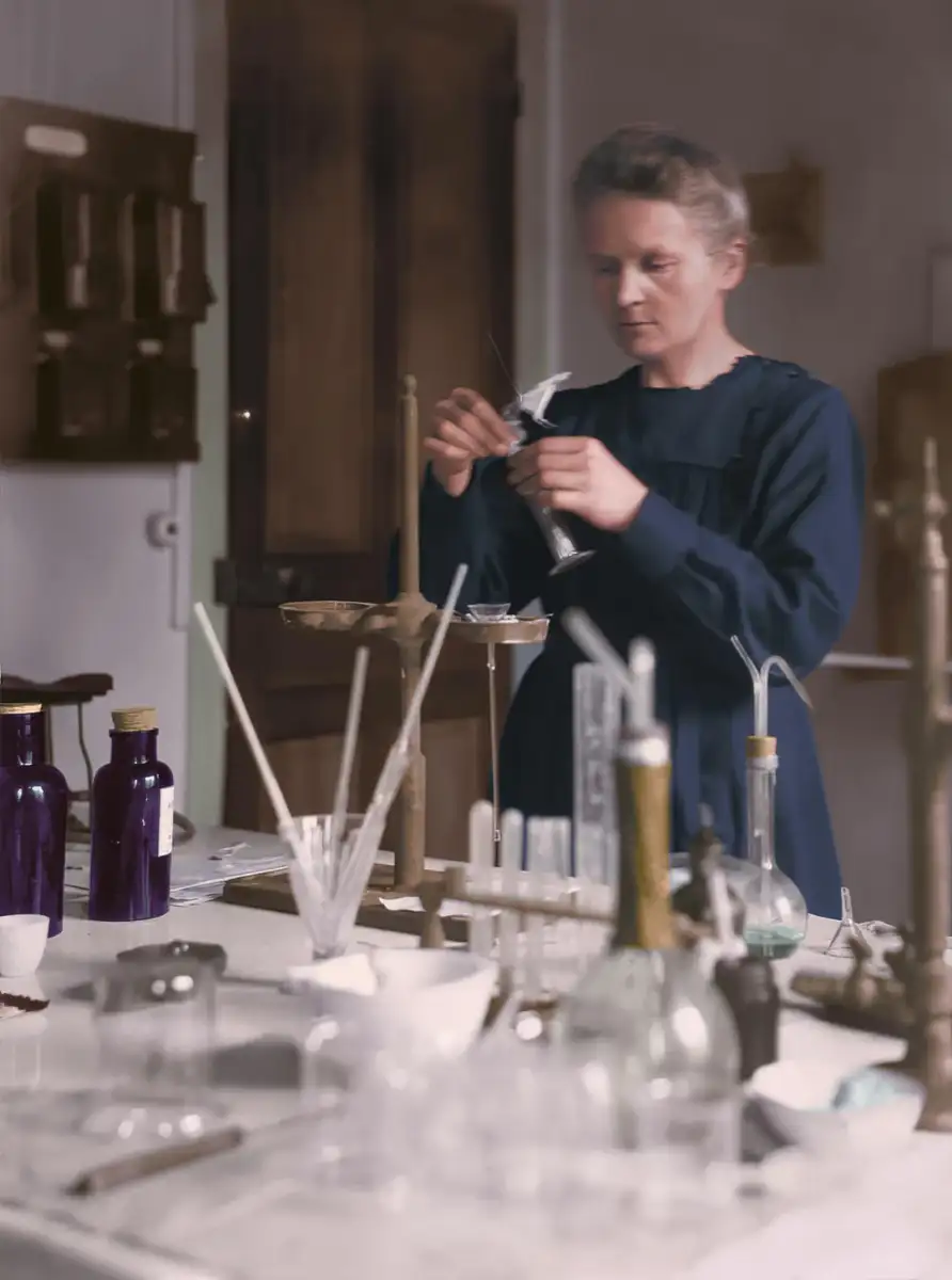 A woman in a dark dress works in a laboratory, handling glassware amid bottles, beakers, and scientific equipment on a cluttered workbench. The setting appears historical.