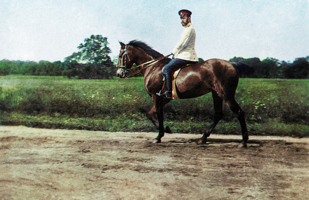 A man in a white military uniform and red hat rides a brown horse along a dirt path, with green grass and trees in the background under a clear sky.