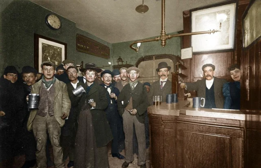 A group of men in early 20th-century clothing stand inside a pub, holding mugs. The interior features wood paneling, a clock, framed pictures, and a bartender behind the counter.