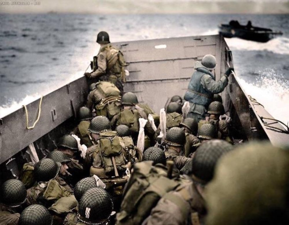 A group of soldiers wearing helmets and gear crouch inside a military landing craft approaching a shore, with waves splashing and another boat visible in the background.