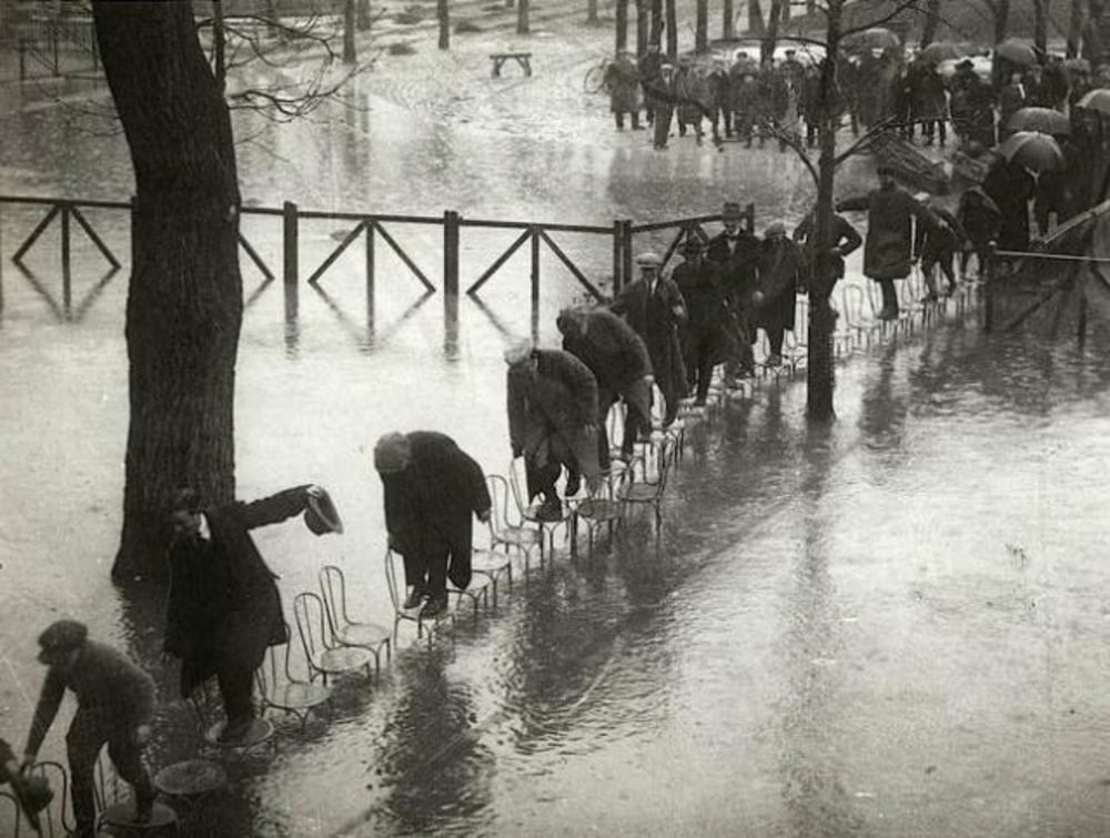 A line of people carefully walk across rows of chairs placed in floodwater, using them as a path to stay dry. Others with umbrellas wait nearby under bare trees and overcast skies.