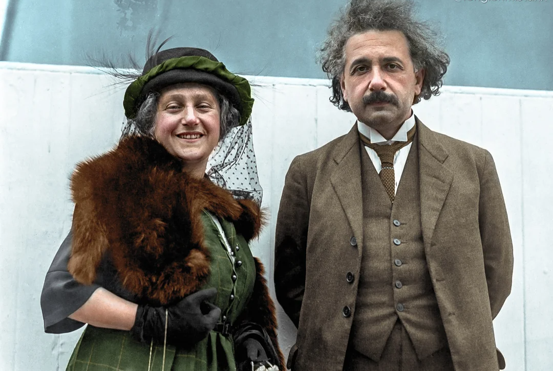 A woman in early 20th-century clothing with a fur stole and hat stands smiling next to a man in a brown suit and tie with wild hair and a mustache. They are posing for a portrait outside.