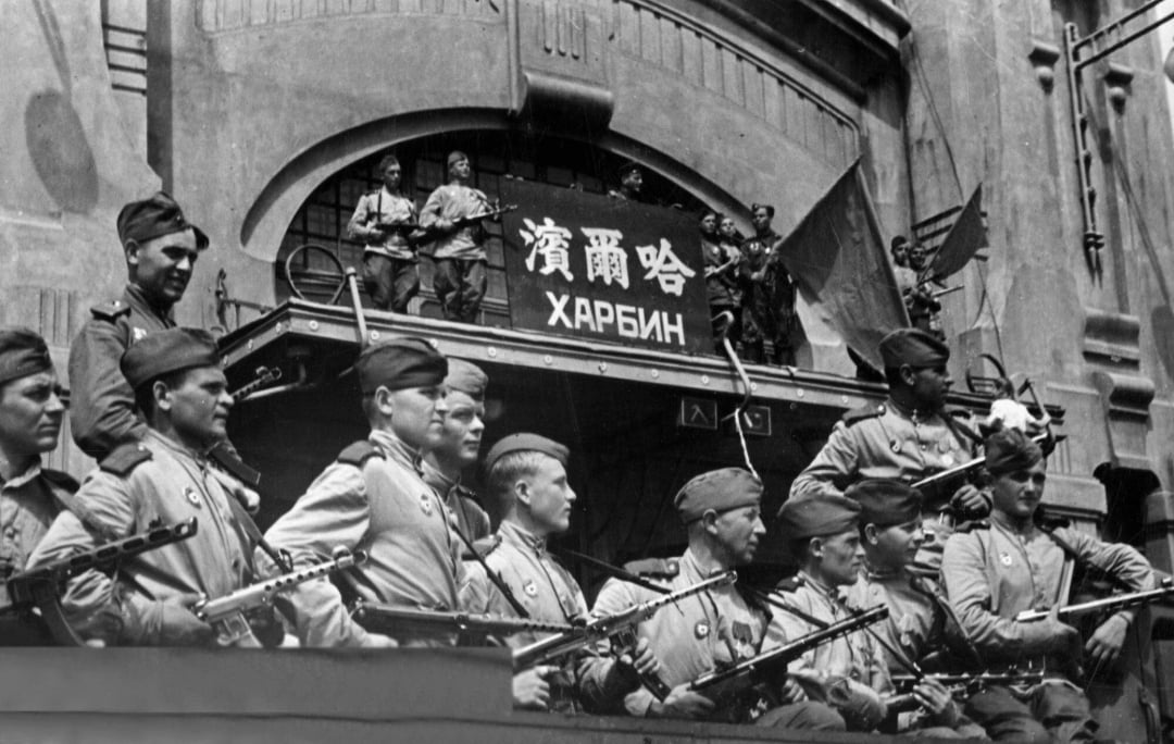 A group of uniformed soldiers with rifles stand and sit in front of a building with a sign reading "Harbin" in Russian and Chinese. More soldiers are visible above, holding weapons and flags. The scene appears historical.