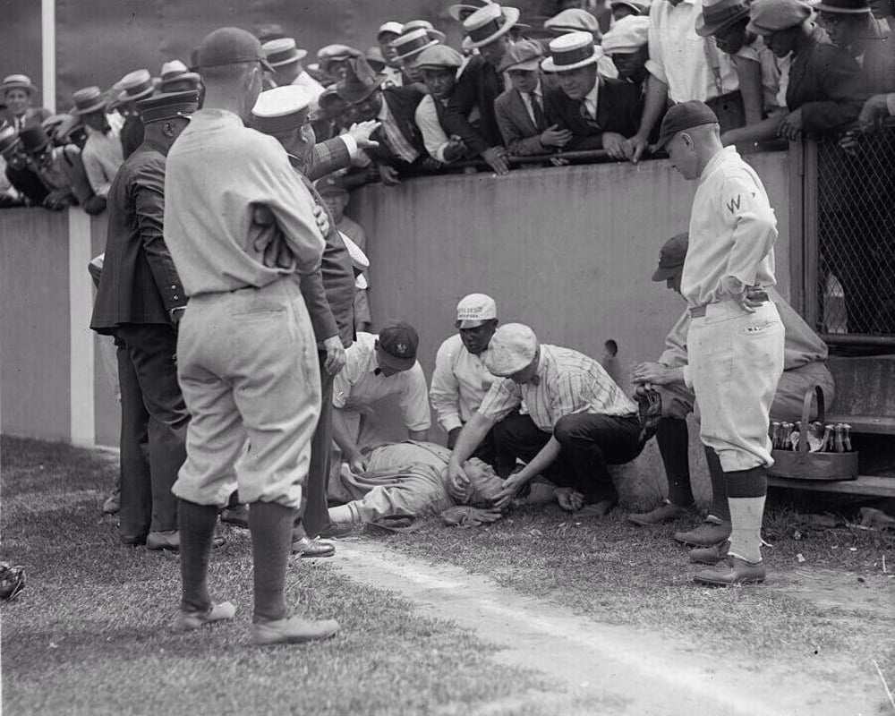 Baseball players and officials gather around an injured player lying on the ground near the field’s edge, while concerned spectators lean over the fence to observe the scene.