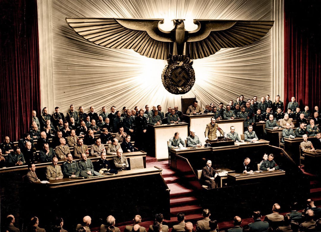 A large assembly hall with many men in military and official uniforms seated, dominated by a large eagle and swastika emblem on the wall behind the speaker’s podium.
