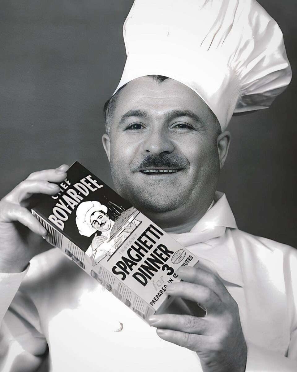 A smiling chef in a traditional white hat and uniform holds a box labeled "Chef Boyardee Spaghetti Dinner" with an illustration of a chef on the packaging. The photo is in black and white.