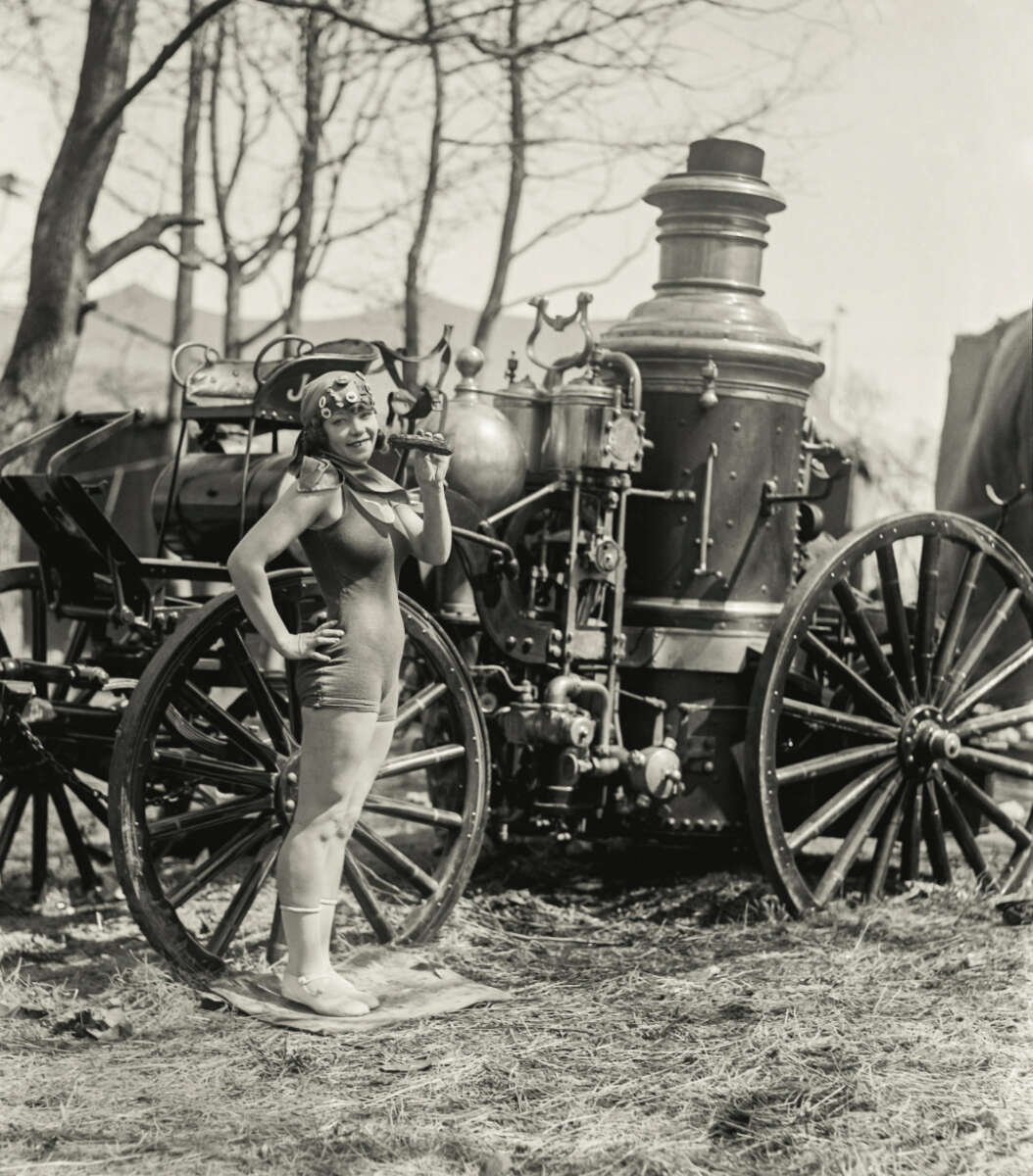 A woman in a vintage swimsuit and swim cap poses confidently in front of a large steam-powered fire engine with tall wheels, outdoors among leafless trees.