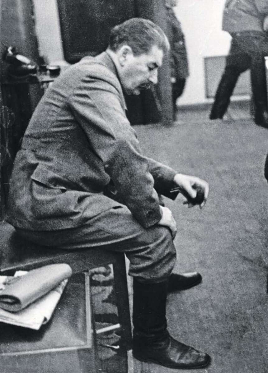 A man in a military-style uniform sits on a bench, looking down with a serious expression. Papers are on the bench beside him, and other people are visible in the background. The image is black and white.
