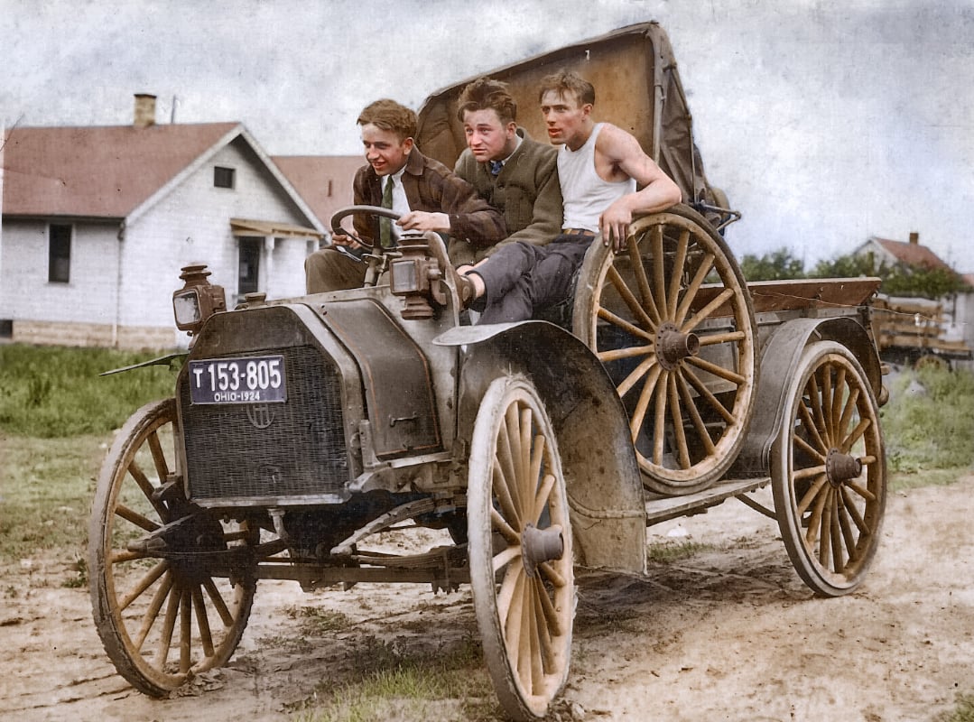 Three young men ride in an old-fashioned, open-top car with large wooden wheels on a dirt road, with houses and grass in the background. The front man steers while the others sit relaxed in the vehicle.