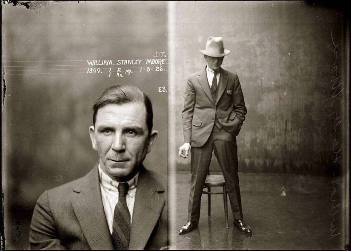 Sepia-toned vintage mugshot featuring a man in a suit and tie. Left: close-up of his face. Right: full-body shot with him standing by a stool, holding a cigarette. Text above reads “WILLIAM STANLEY MOORE” with ID numbers and a date.