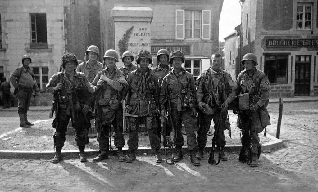A group of World War II soldiers in full gear stands together in a European village square, with buildings and a memorial behind them. The soldiers look worn but resolute, and their uniforms are heavy with equipment.