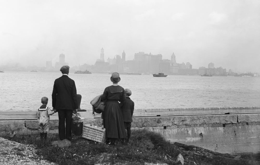 A family of four, dressed in early 1900s clothing, stands on a shoreline with suitcases, looking across the water at the distant, hazy skyline of New York City.