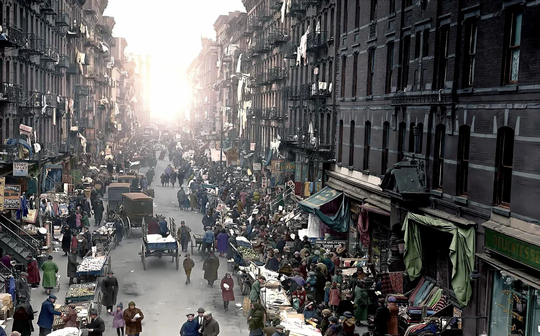 A bustling city street market scene with crowds of people, street vendors, carts, and market stalls lining both sides of the road between tall historic buildings; sunlight shines at the end of the street.